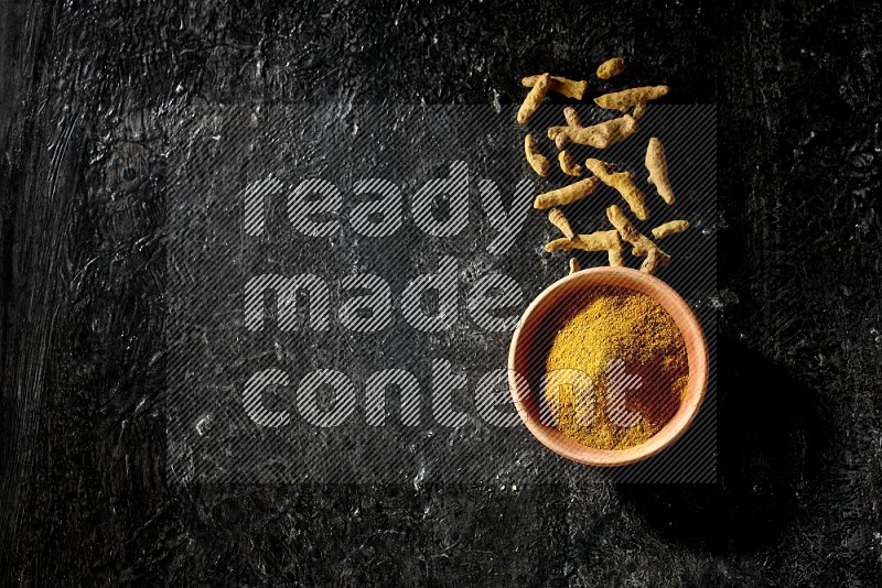 A wooden bowl full of turmeric powder with dried turmeric fingers on textured black flooring