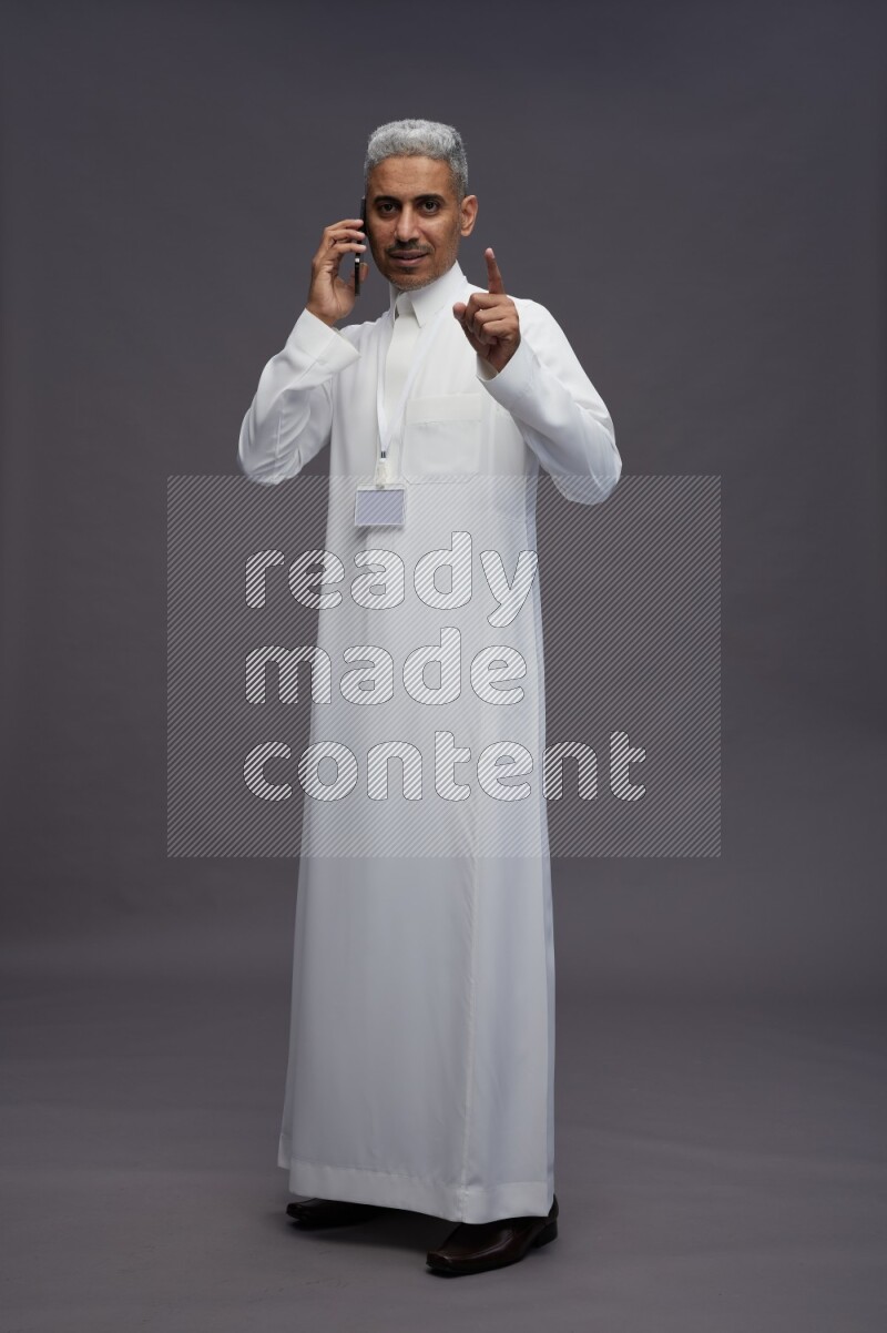 Saudi man wearing thob with neck strap employee badge standing talking on phone on gray background