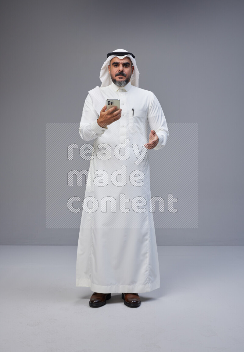 Saudi man Wearing Thob and white Shomag standing texting on phone on Gray background