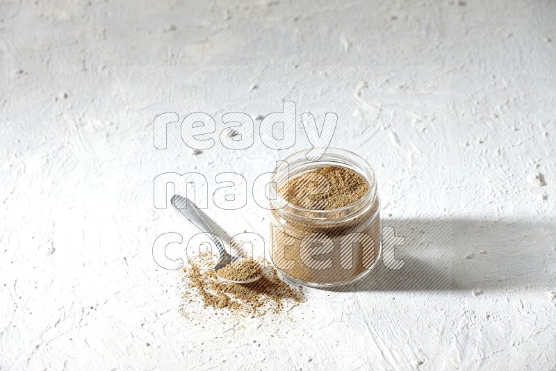 A glass jar and a metal spoon full of cumin powder on textured white flooring