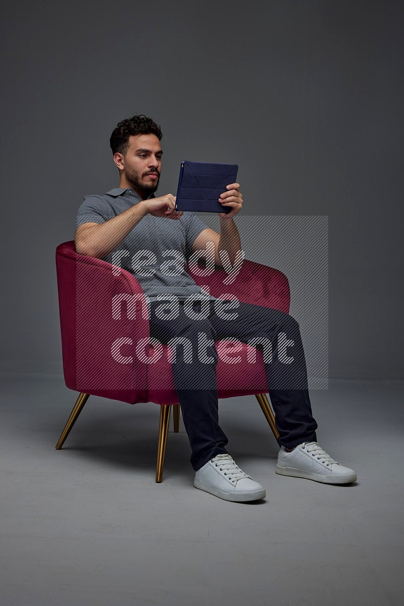 A man wearing casual and using his tablet while sitting on a burgundy chair eye level on a gray background