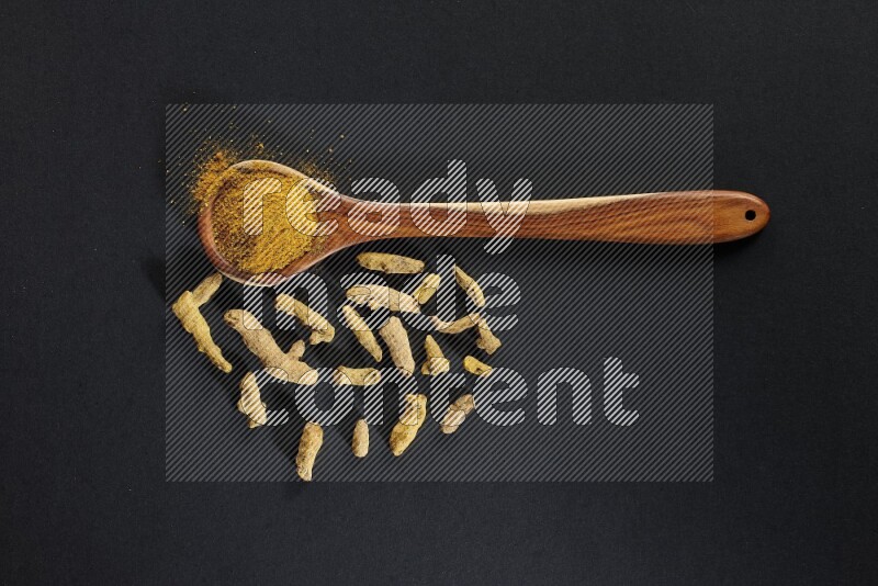 A wooden ladle full of turmeric powder with dried turmeric fingers on black flooring