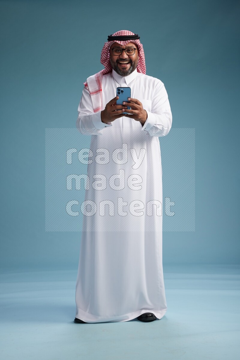 Saudi Man with shimag Standing texting on phone on blue background