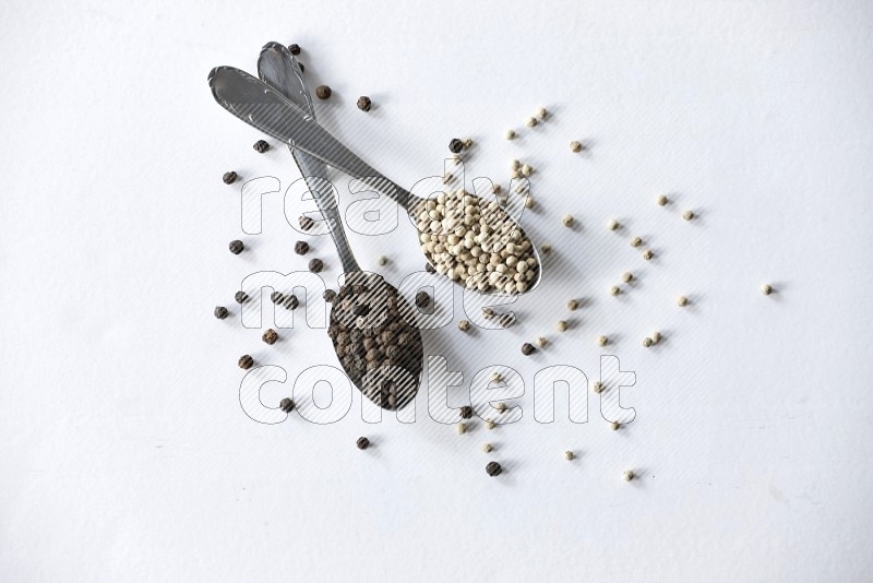 2 metal spoons full of black and white pepper beads with spreaded beads on white flooring