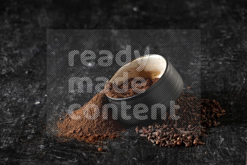 وعاء فخاري أسود ممتلئ ببودرة القرنفل والبودرة تناثرت منه مع الحبوب على خلفية سوداء