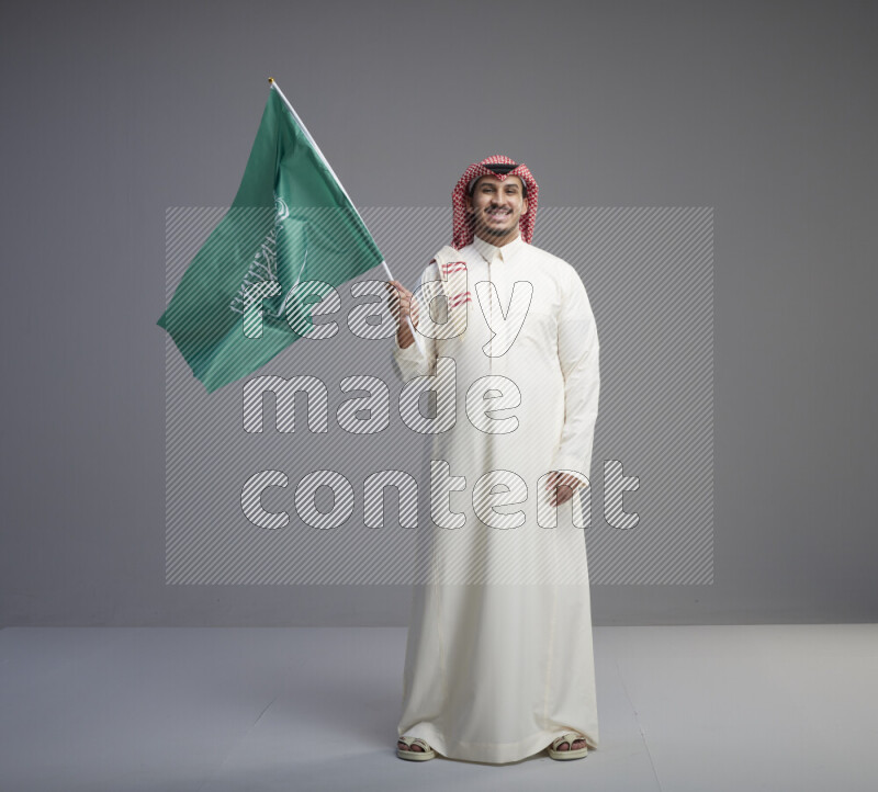 A Saudi man standing wearing thob and red shomag raising big Saudi flag on gray background