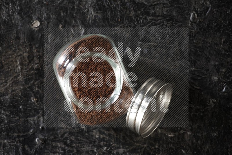 A glass spice jar full of cloves powder on textured black flooring