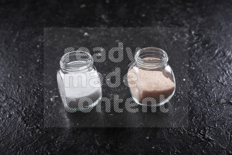 2 glass jars one is filled with fine himalayan salt and the other with table salt on black background