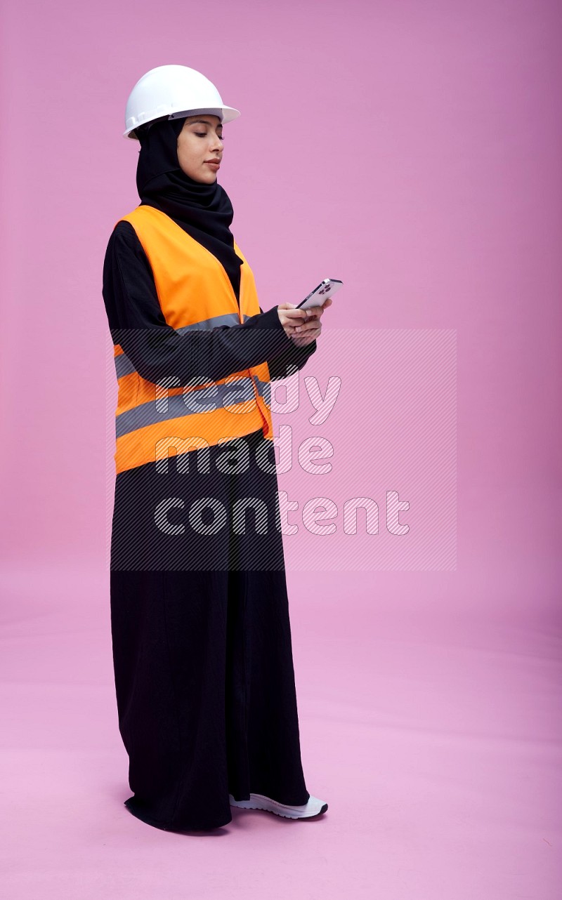 Saudi woman wearing Abaya with engineer vest and helmet standing texting on phone on pink background