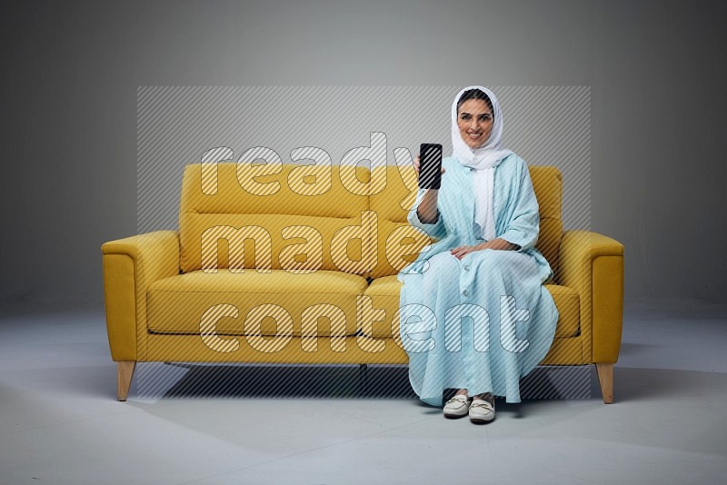A Saudi woman wearing a light blue Abaya and white head scarf sitting on a yellow sofa and showing her phone's screen vertically and horizontally eye level on a grey background