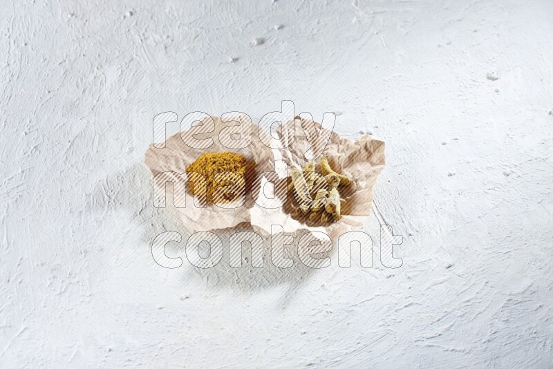 Turmeric powder and dried turmeric whole fingers in 2 crumpled pieces of paper on textured white flooring