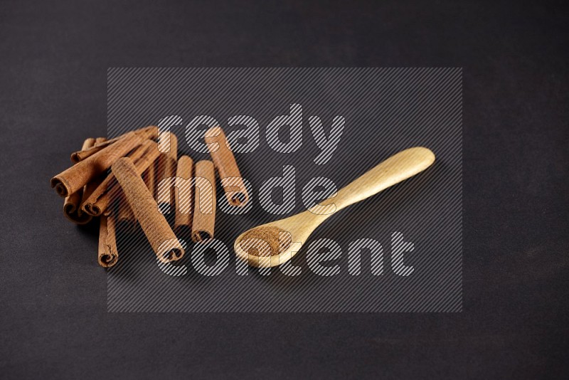 Cinnamon sticks stacked beside a wooden spoon full of cinnamon powder on black background