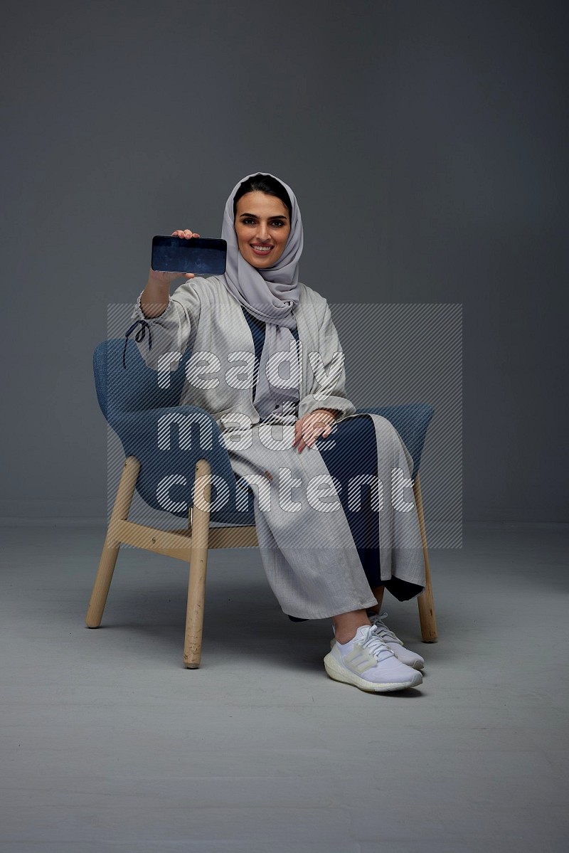 A Saudi woman wearing a light gray Abaya and head scarf sitting on a dark grey chair and showing the phone's screen eye level on a grey background