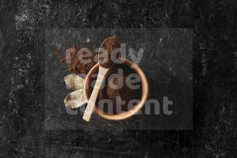 A wooden bowl and a wooden spoon full of cloves powder with laurel leaves on a textured black flooring