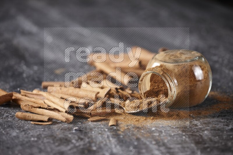 Herbal glass jar full cinnamon powder flipped and a metal spoon full of powder surrounded by cinnamon sticks on textured black background in different angles