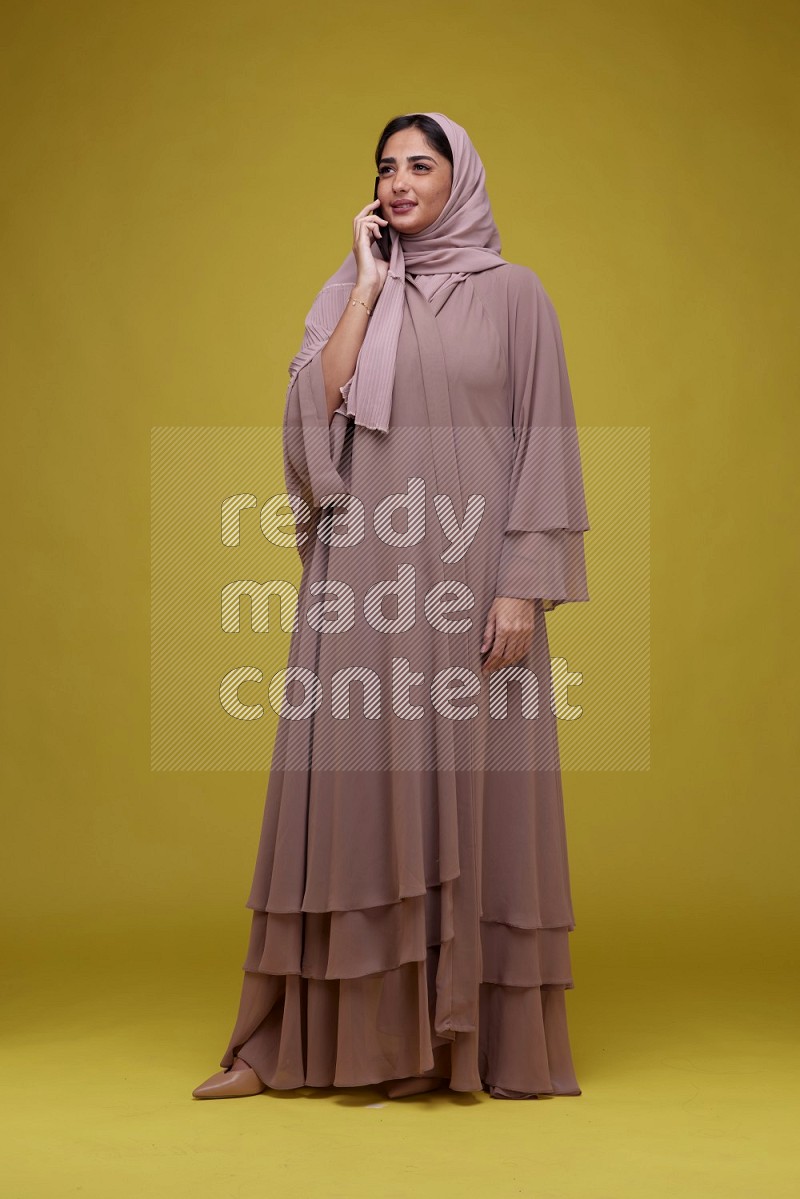 A woman having a call on a Yellow Background wearing Brown Abaya with Hijab