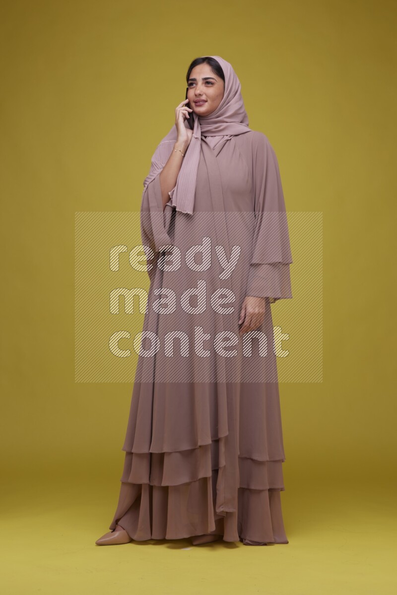 A woman having a call on a Yellow Background wearing Brown Abaya with Hijab