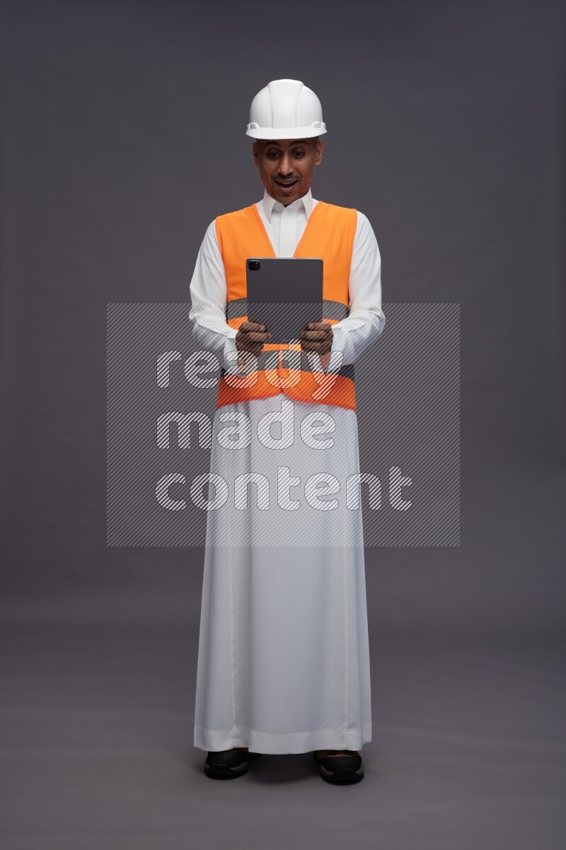 Saudi man wearing thob with engineer vest standing working on tablet on gray background