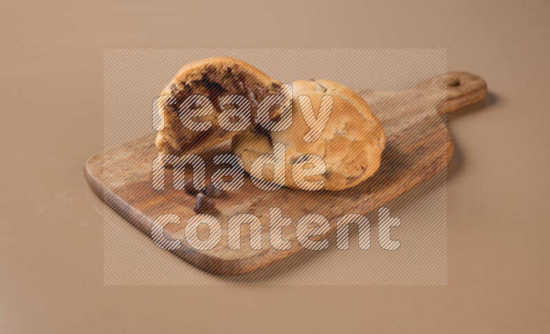 a chocolate chip cookie with another one cut in half on a wooden cutting board on a brown background