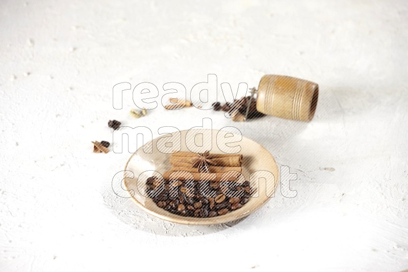 Beige plate full of coffee beans, cinnamon sticks and star anise with a coffee grinder, coffee beans, cinnamon pieces and cardamom next of it on white background