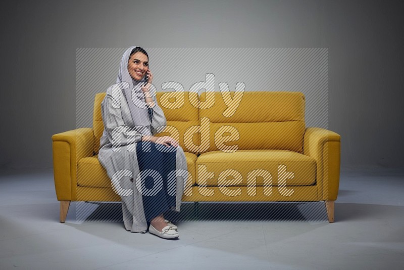 A Saudi woman wearing a light gray Abaya and head scarf sitting on a yellow sofa and talking in the phone eye level on a grey background