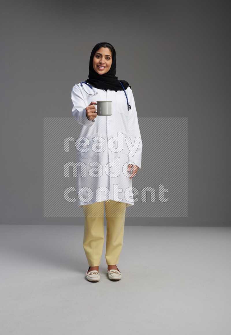 Saudi woman wearing lab coat with stethoscope standing holding mug on Gray background
