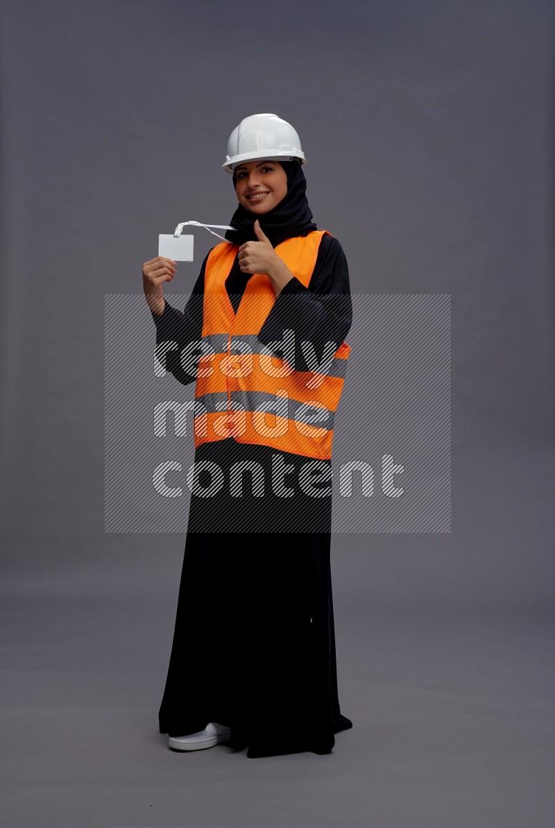 Saudi woman wearing Abaya with engineer vest with neck strap employee badge standing interacting with the camera on gray background