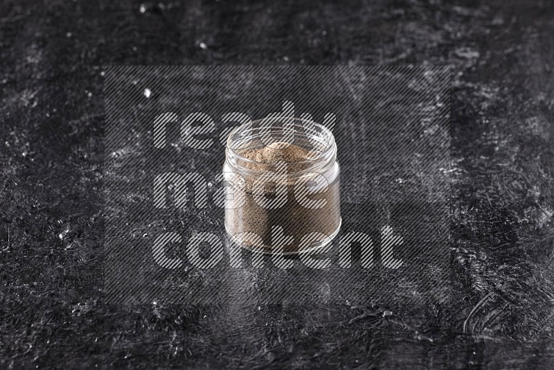A glass jar full of black pepper powder on a textured black flooring