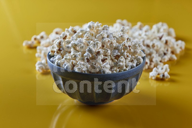 A blue pottery bowl full of popcorn with popcorn beside it on a yellow background in different angles