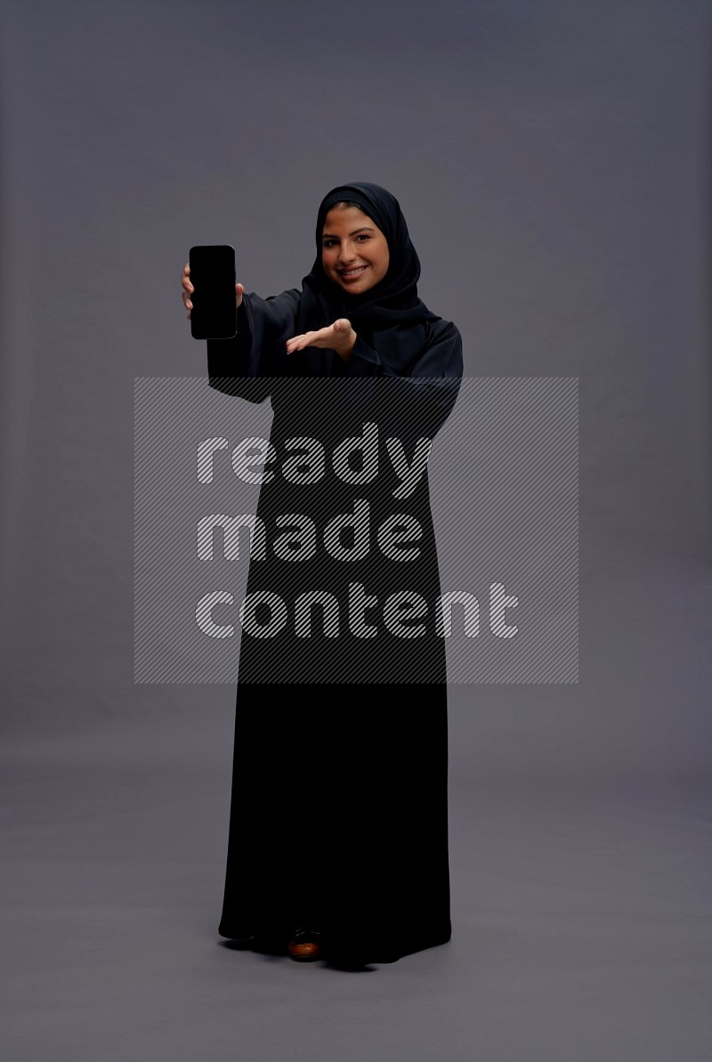 Saudi woman wearing Abaya standing showing phone to camera on gray background