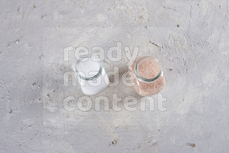 2 glass jars one is full of fine himalayan salt and the other with table salt on white background