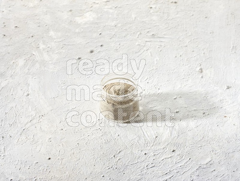 A glass jar full of white pepper powder on textured white flooring