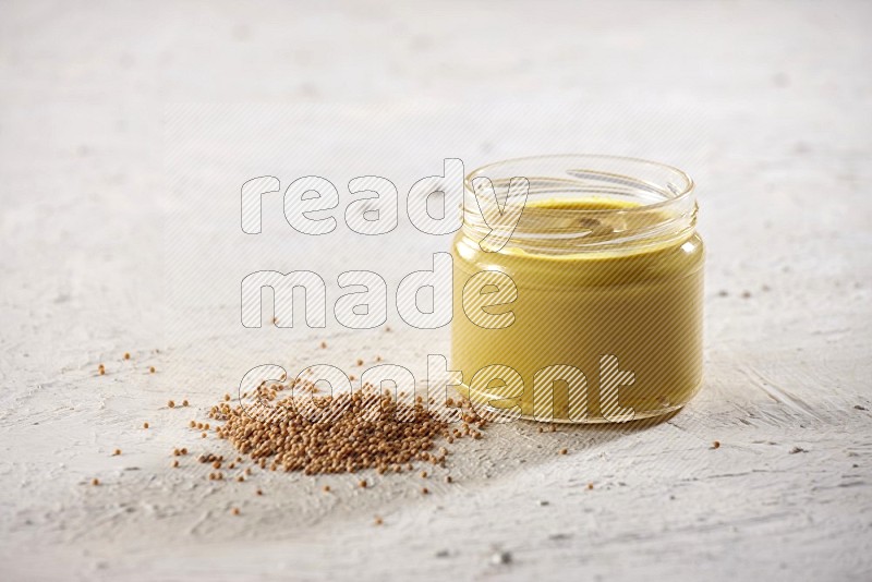 A glass jar full of mustard paste with mustard seeds spread next to it on a textured white flooring
