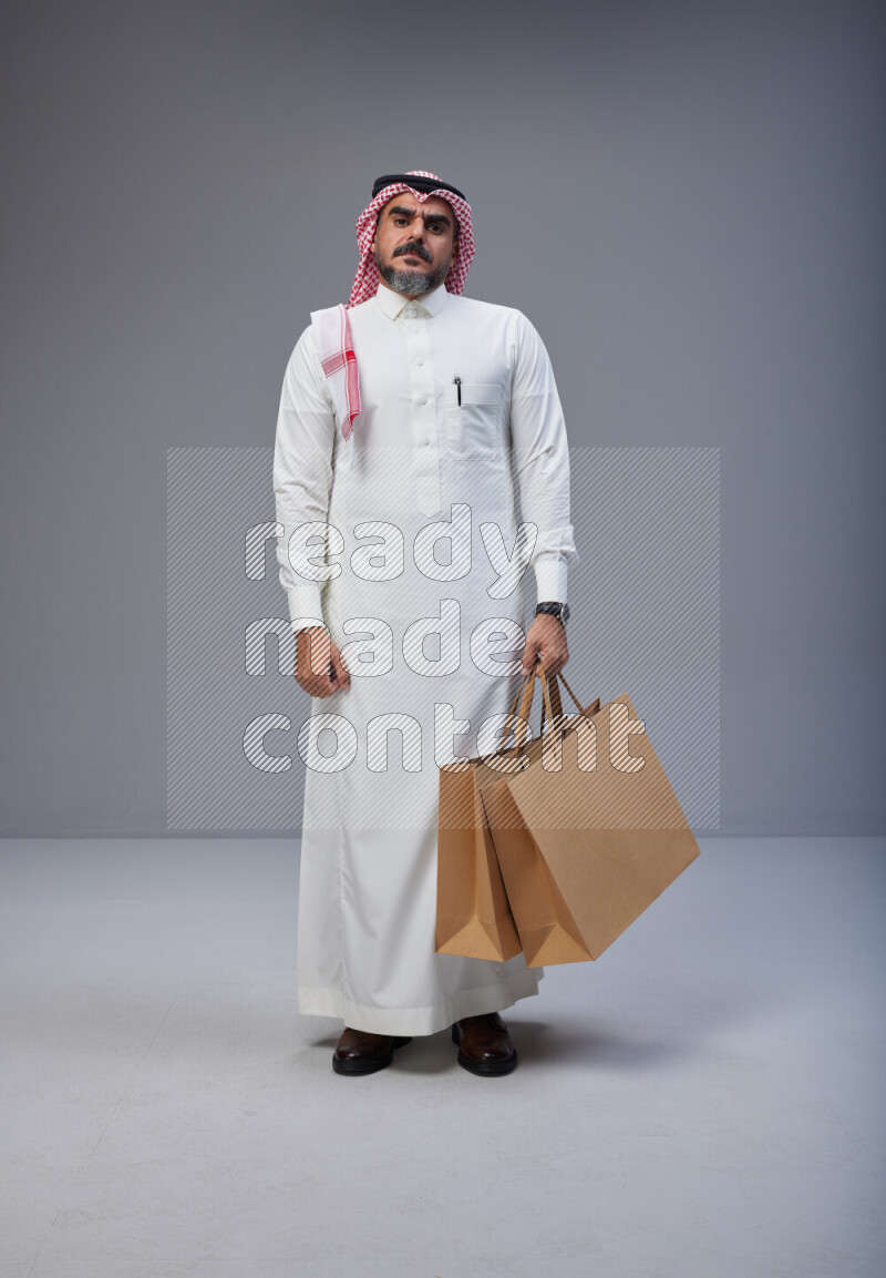 Saudi man Wearing Thob and red Shomag standing holding shopping bag on Gray background