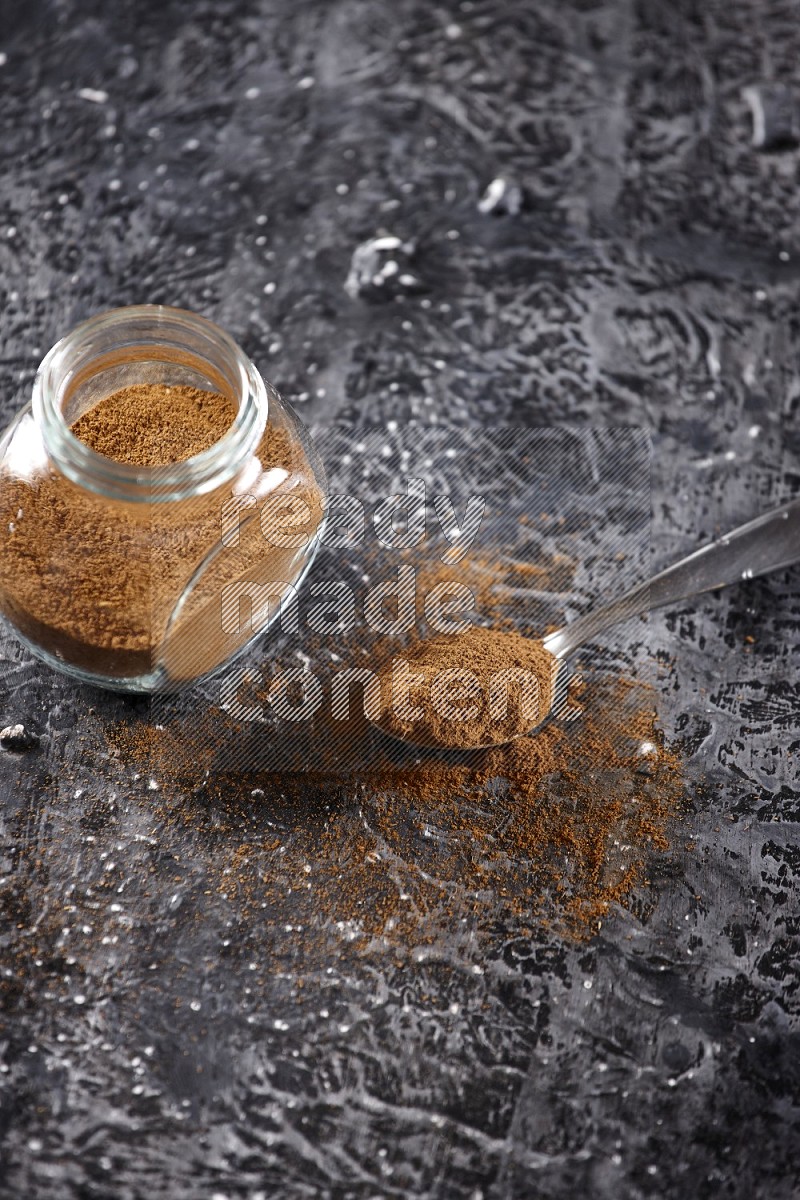 Herbal glass jar full of cinnamon powder and a metal spoon full of powder on textured black background