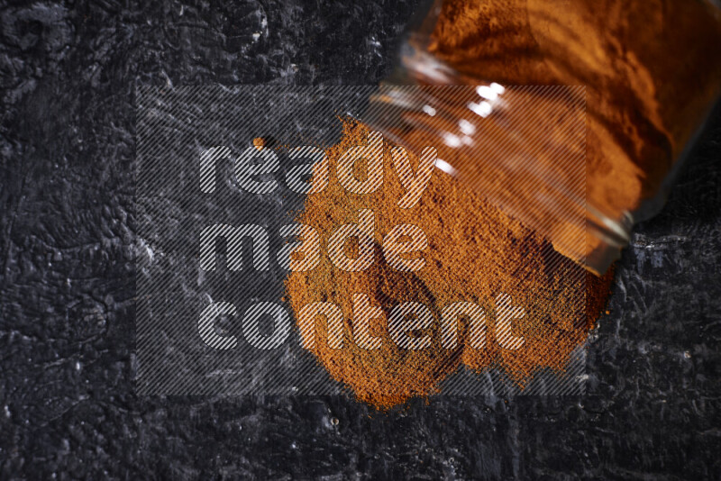 A glass jar full of ground paprika powder flipped with some spilling powder on black background