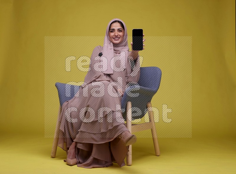 A woman Showing Her Phone Screen Setting  on a Yellow Background wearing Brown Abaya with Hijab