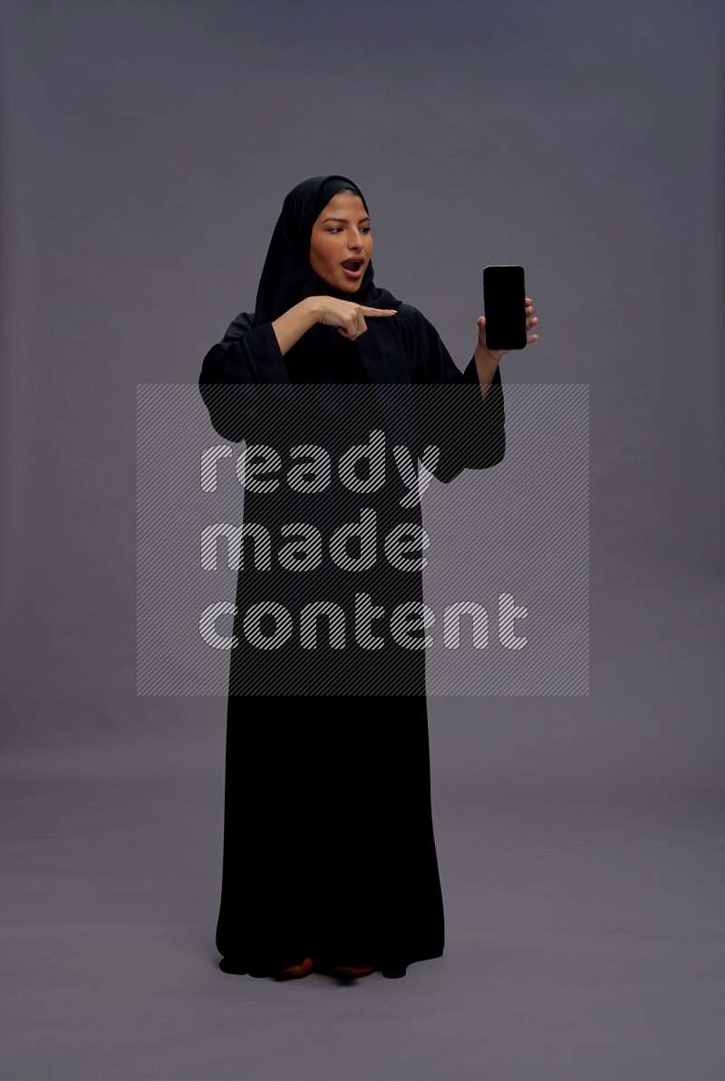 Saudi woman wearing Abaya standing showing phone to camera on gray background