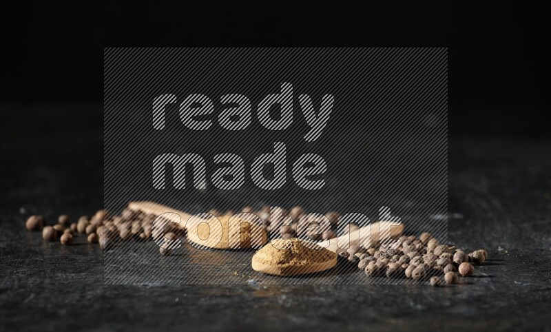 Wooden spoons full of all spice powder and allspice whole balls beside it on a textured black flooring