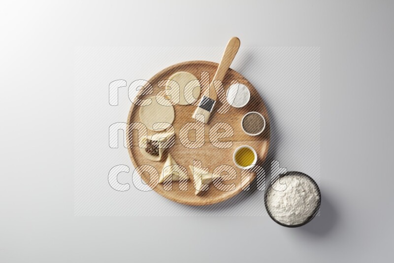 two closed sambosas and one open sambosa filled with meat while flour, salt, black pepper and oil with oil brush aside in a wooden dish on a white background