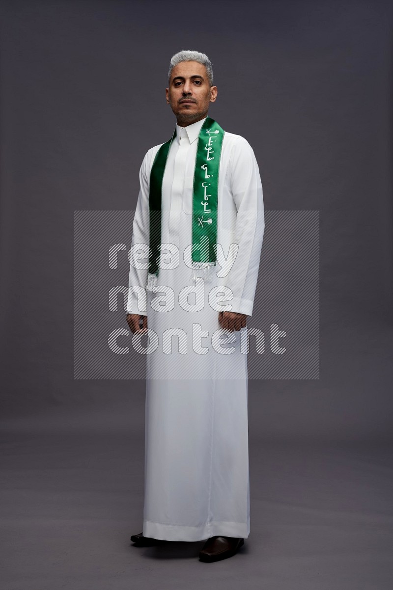 Saudi man wearing thob standing holding Saudi flag on gray background