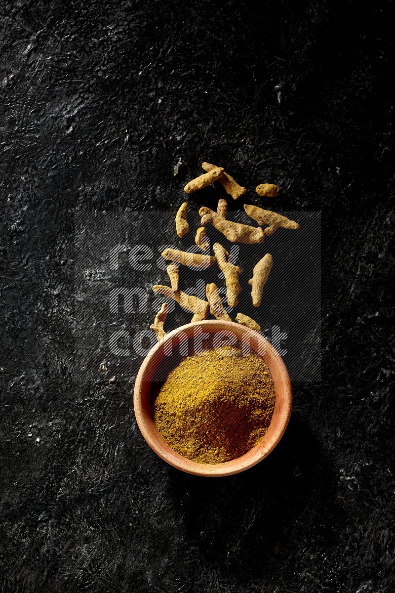 A wooden bowl full of turmeric powder with dried turmeric fingers on textured black flooring