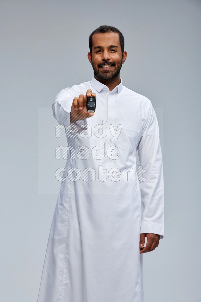 Saudi man wearing thob and shomag standing holding car key on gray background