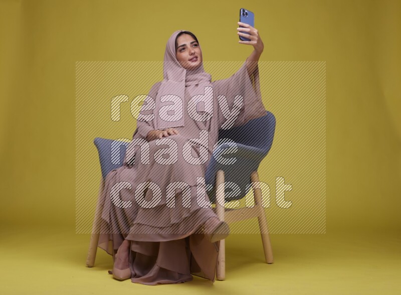 A woman sitting on a blue chair and Taking a Selfie  Setting  on a Yellow Background wearing Brown Abaya with Hijab