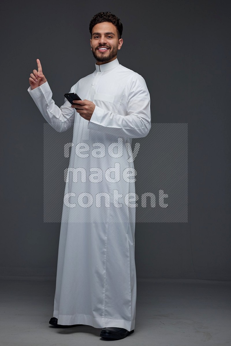 A Saudi man wearing Thobe and holding his phone while pointing to it eye level on a gray background