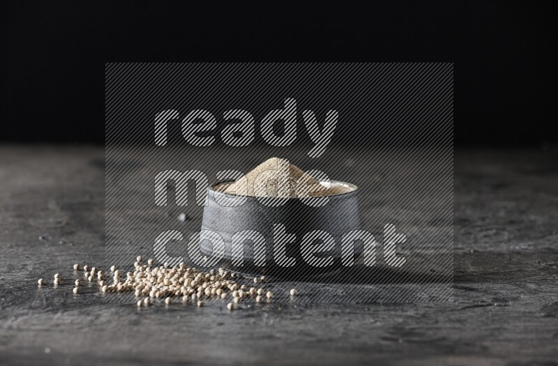 Black pottery bowl full of white pepper powder with white pepper beads on textured black flooring