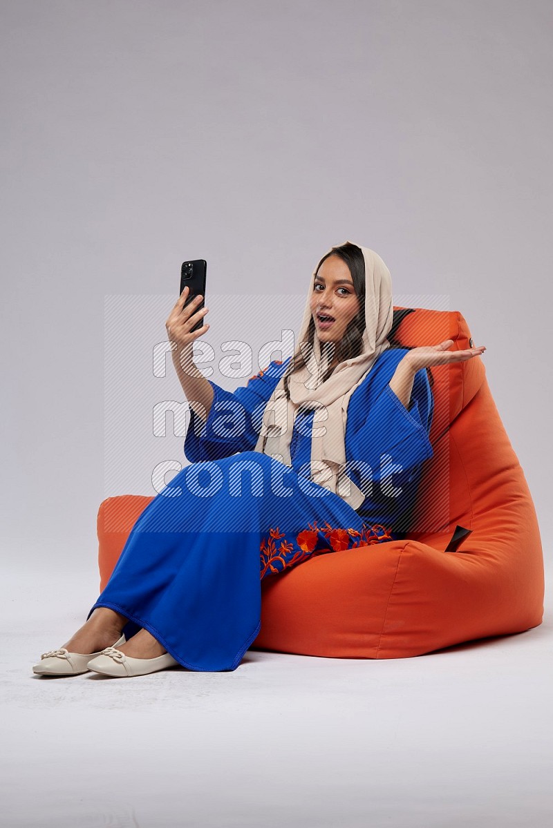 A Saudi woman wearing jalabiya sitting on an orange beanbag and taking selfie