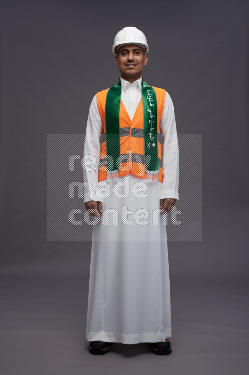 Saudi man wearing thob with engineer vest standing holding Saudi flag on gray background