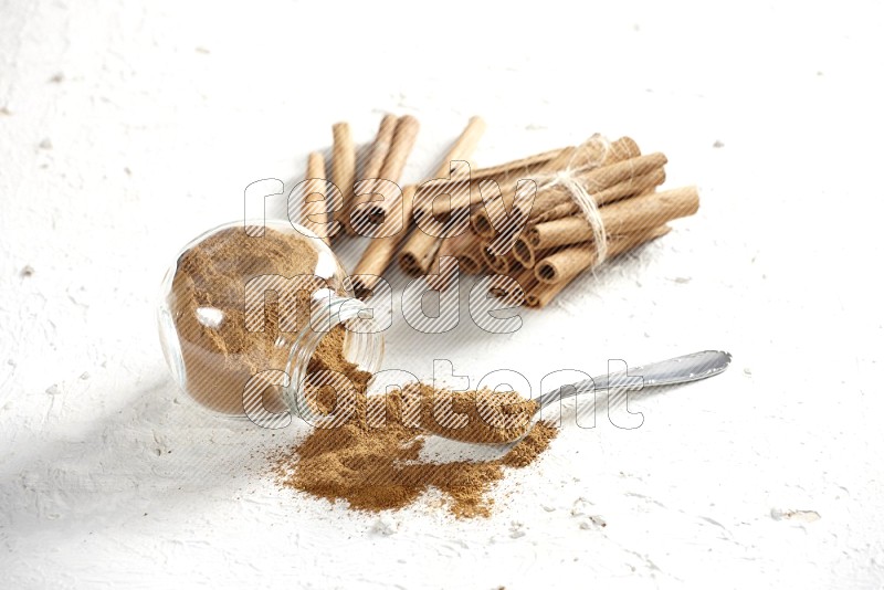 Flipped herbs glass jar full of cinnamon powder with a metal spoon full of powder and cinnamon sticks on a textured white background