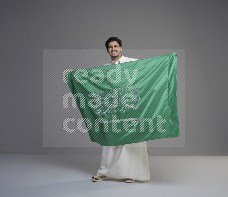 A Saudi man standing wearing thob holding big Saudi flag on gray background